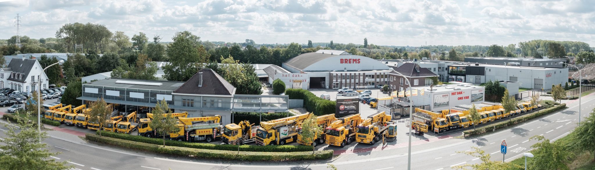 Wagenpark van Brems Dakwerken uit Vilvoorde, met een rij gele hoogwerkers op vrachtwagen opgesteld voor hun bedrijfsgebouw
